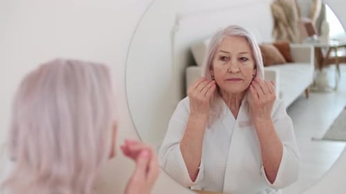 Senior Woman Applies Moisturizer in Front of Mirror
