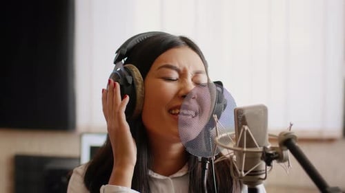 Asiatische Musikerin mit Kopfhörern singt Lied zum Mikrofon im Musikstudio