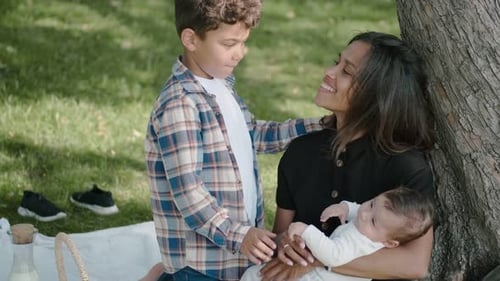 Family Picnic in the Summer Park. A Mother with Two Sons Is Sitting Near a Tree, a Boy Kisses His