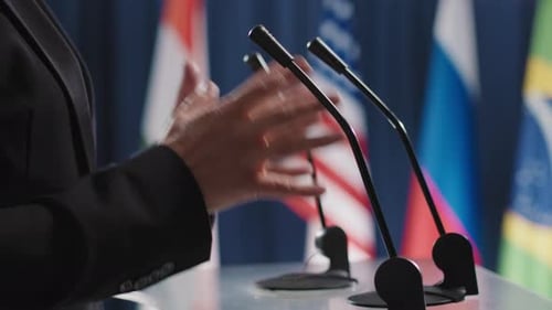Adult Gesturing at Podium with Flags in Background
