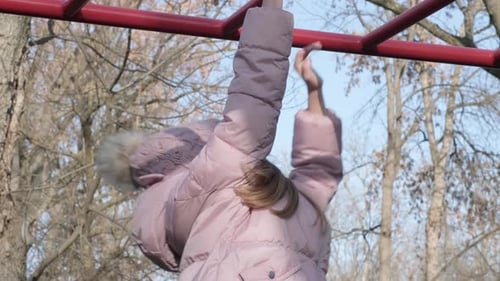 Side View of Little Girl Climbs on Horizontal Bars on Playground in Autumn Park