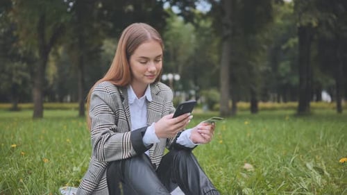 A Young Girl Makes a Purchase with Her Phone and Bank Card in the Park Sitting on the Grass