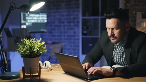 Male Employee Using Laptop Working Overtime at Night in Office Busy with Project