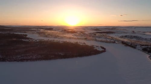 Aerial Over the Frozen Winding River Surrounded with Forest and with a Little Village on a Side