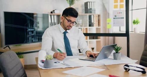 Trabajador barbudo con gafas que escribe notas sobre un plan comparando datos con información en una computadora portátil