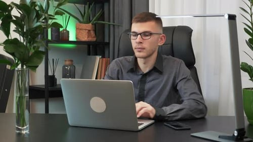 Young man is closing laptop computer and finishing to work at workplace in office in the evening
