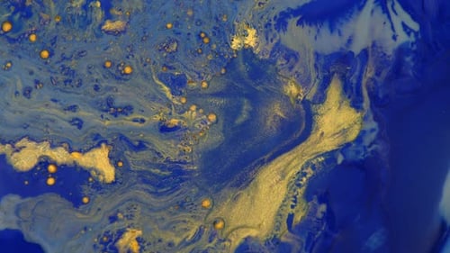 Process of mixing liquid blue and gold paint. Floating spots of golden paint mix with blue color.