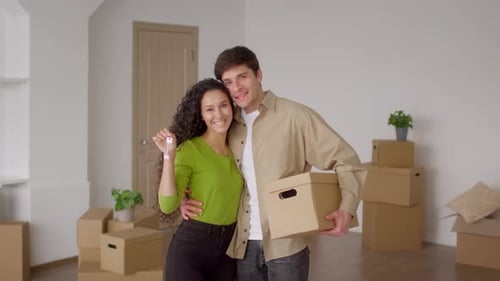 Spouses Holding New House Key Embracing Carrying Moving Box Indoor