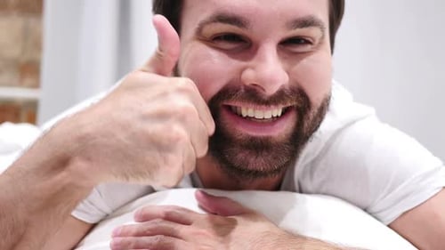 Pouces vers le haut par un homme à barbe heureux allongé dans son lit sur le ventre