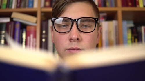 A Young Man Is Reading a Book in a Library. A Man with Glasses Carefully Looks at the Book Closeup
