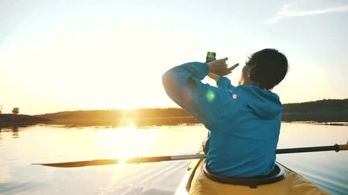Man Makes Selfie on Smartphone While Sitting in Kayak on Calm Lake at Sunset, Outdoor Activities