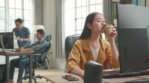 Asian Female Drinking Coffee While Works On Her Personal Computer With Big Display In Office
