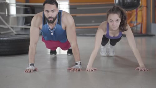 Front View of Young Caucasian Woman and Middle Eastern Man Pushing Up in Gym