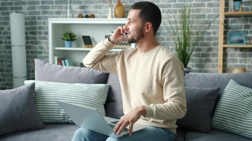 Man Using Laptop and Talking on Phone at Home