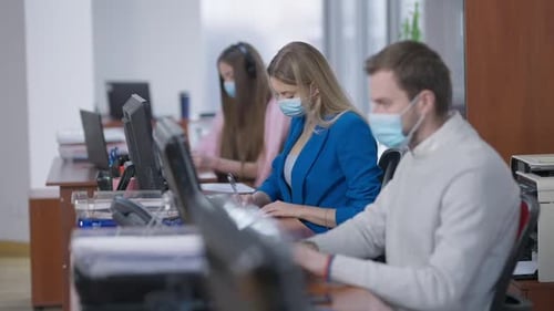 Side View of Concentrated Blond Young Woman in Covid Face Mask Working in Office with Blurred