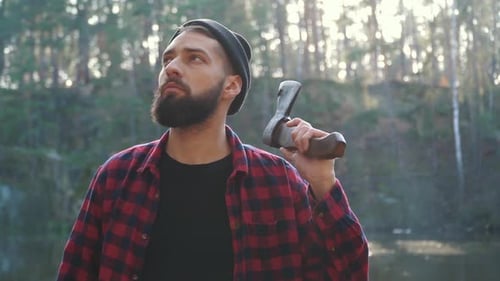 Young Bearded Man with an Axe Walk in Forest. Unshaven Forester with an Ax Outdoors