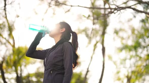 4K Asian woman drinking water from a bottle while jogging at public park in the morning.