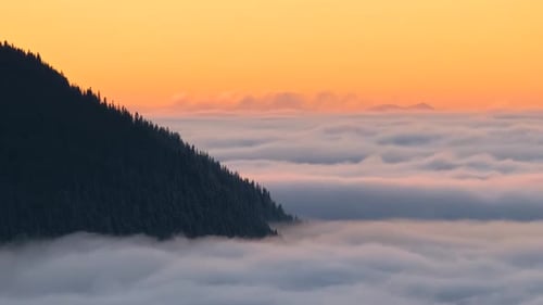 Time lapse of distant dark mountain hills surrounded with white fast moving foggy clouds at sunrise.