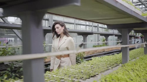 Attractive Young Caucasian Woman Walking in Glasshouse Along Rows of Green Plants and Flowers