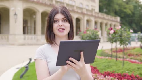 A Young Caucasian Woman Works on a Tablet and Talks to the Camera with a Smile in a City Park