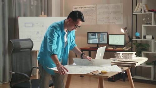 Asian Man Engineer With A Laptop Looking At Blueprint While Working At The Office