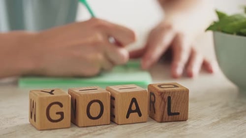 Das Wort Goal ist aus Holzwürfeln zusammengesetzt. Im Hintergrund schreibt eine weibliche Hand das Ziel
auf