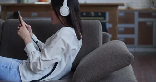 Glückliche asiatische Frau, die Songs mit Kopfhörern hört und Smartphone berührt Bildschirm auf gemütlichem