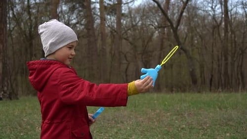 A Boy in a Red Jacket and Hat Blows Soap Bubbles on the Street. A Child Plays in the Fresh Air in