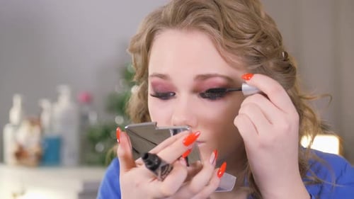 Augen-Make-up. Das Mädchen sieht in einem kleinen Handspiegel aus. Tragen Sie schwarze Farbe auf die Wimpern
auf