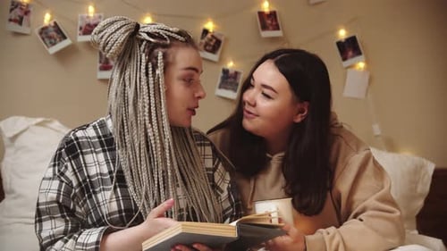 Young Women Reading Book Together Indoors in Bed