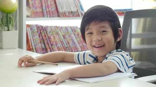 Little Asian Child Using A Pencil To Write On Notebook At The Desk