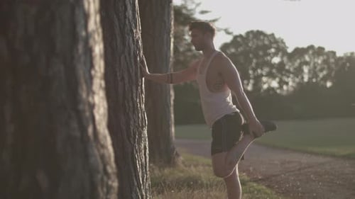Man In Park Stretching Out His Legs Before He Goes For a Run Whilst Sun Sets Behind Him In Slow Moti