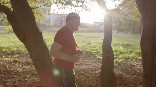 Male Senior Person Running Along the Road in Park. Mature Runner Man Training, Listening Music