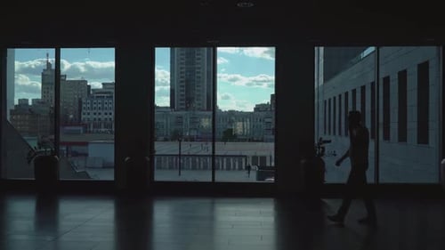 Man Goes Dark Corridor of Office Space with Large Windows.