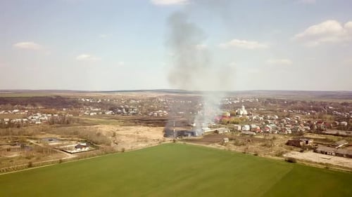Aerial view of a field with dry grass set on fire with orange flames and high column of smoke.