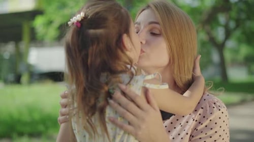 Close-up Portrait of Beautiful Young Caucasian Mother Kissing Pretty Daughter in Sunny Park. Smiling
