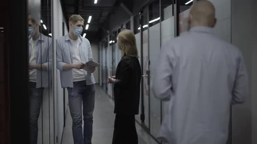 Concentrated Young Caucasian Man and Woman in Covid Face Masks Standing in Office Corridor Talking