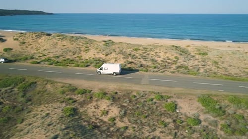Drone Follows White Van or Minibus Driving Along Coastal Road Next To Sand Beach
