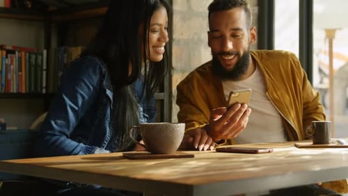 Front view of happy young mixed-race couple discussing over mobile phone at table in cafe 4k