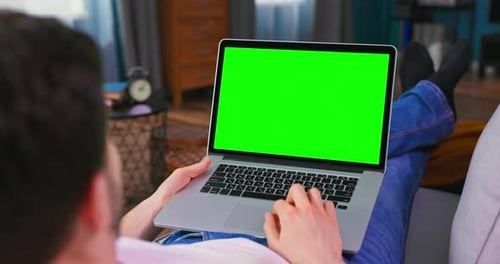 Man at Home Lying on a Couch Working on Laptop Computer with Green Mockup Screen