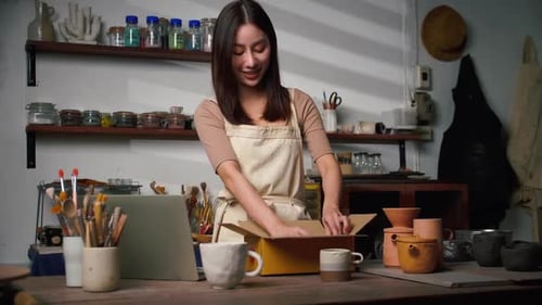 A young Asian woman ceramist is packing ceramic mugs in a parcel box for people who buy online.