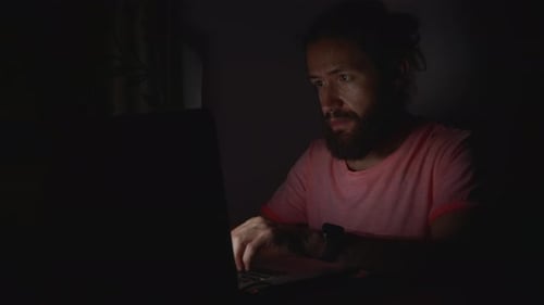 Junger bärtiger Mann arbeitet im Dunkeln am Laptop