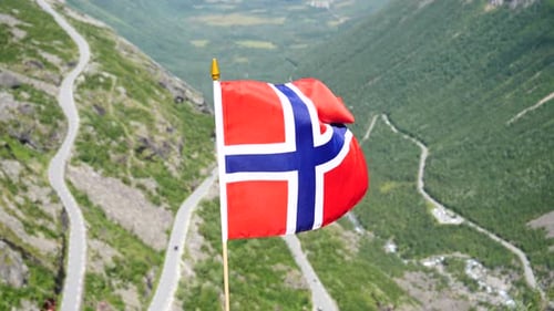 Norwegian Flag Waving Above Winding Mountain Road
