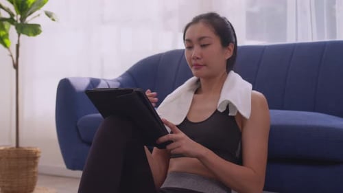 Asiatin in Sportbekleidung wischt sich die Stirn mit Handtuch ab und benutzt digitales Tablet, während sie sich nach zwei ausruht