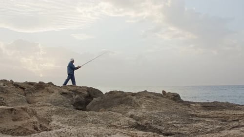 Old fisherman standing on sea side rocks and fishing against the sunset
