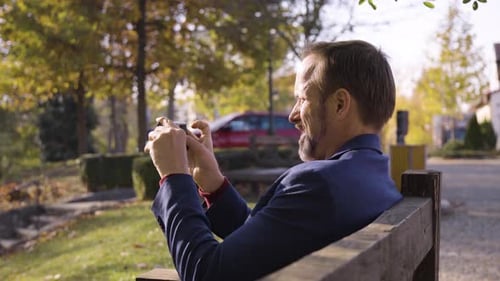 Man Uses Smartphone on Park Bench on Sunny Day