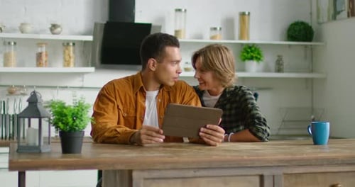 Happy Mixed Race Couple Sitting in Their Kitchen, Using Tablet Computer