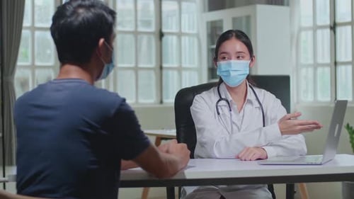 Woman Doctor Talking With Male Patient During Consultation In A Health Clinic. Both Wear Face Masks
