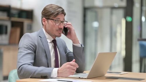 Businessman with Laptop Talking on Smartphone in Office
