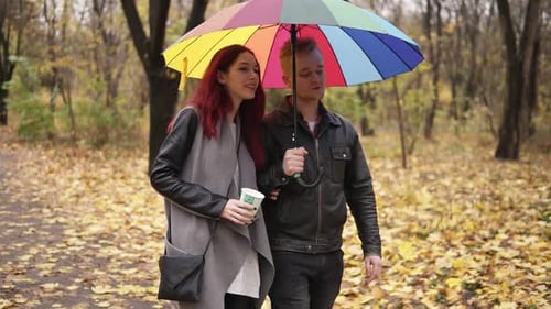 Junges glückliches Paar geht zusammen im Herbstpark spazieren und hält einen bunten Regenschirm